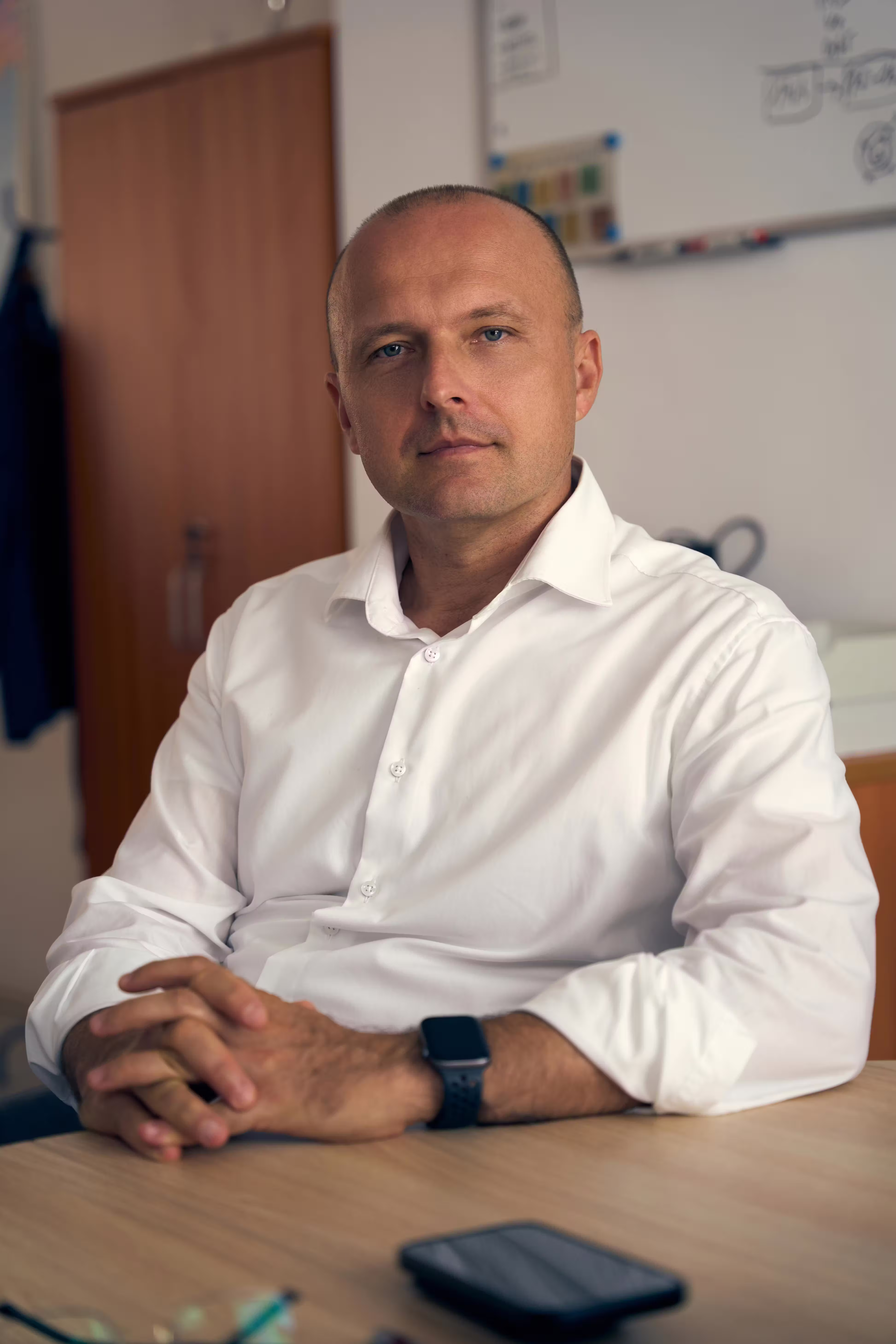Man in a white shirt sitting at a desk with hands clasped, looking at the camera.