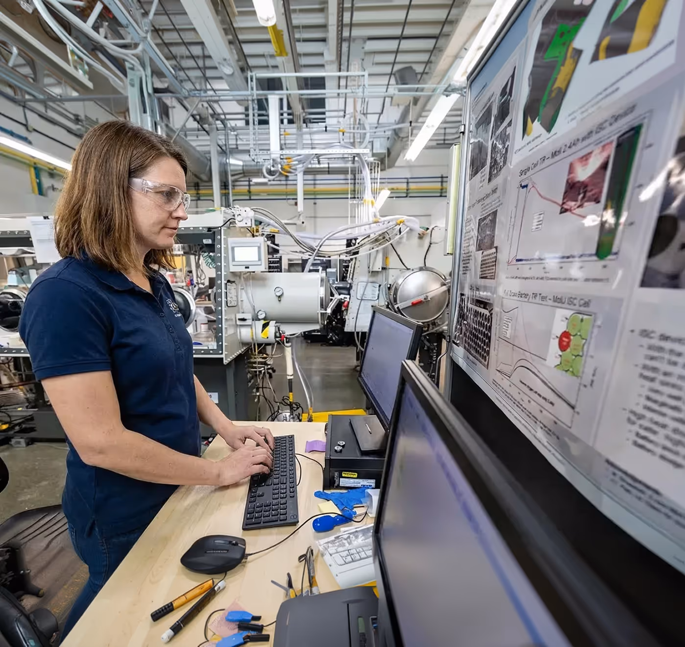 Woman wearing safety glasses typing on a keyboard at a workstation with multiple monitors in an industrial laboratory.