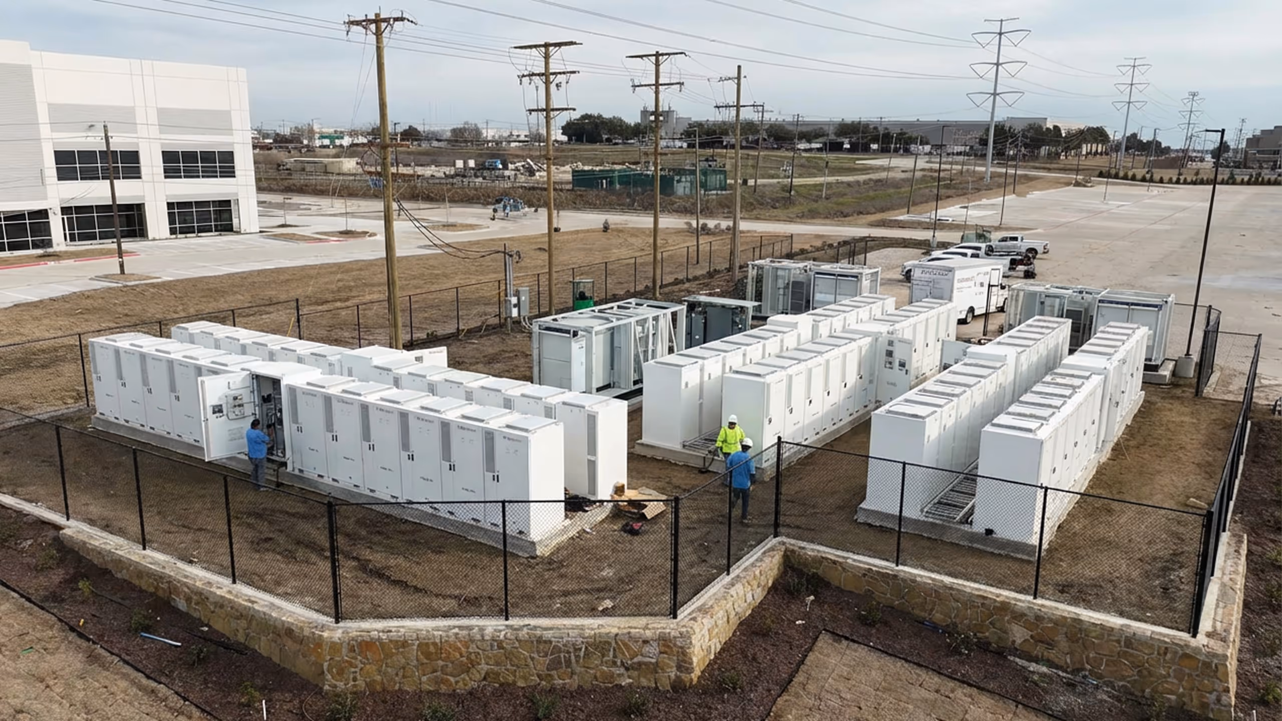 Outdoor energy storage facility with rows of large white electrical cabinets and three workers inside a fenced area.