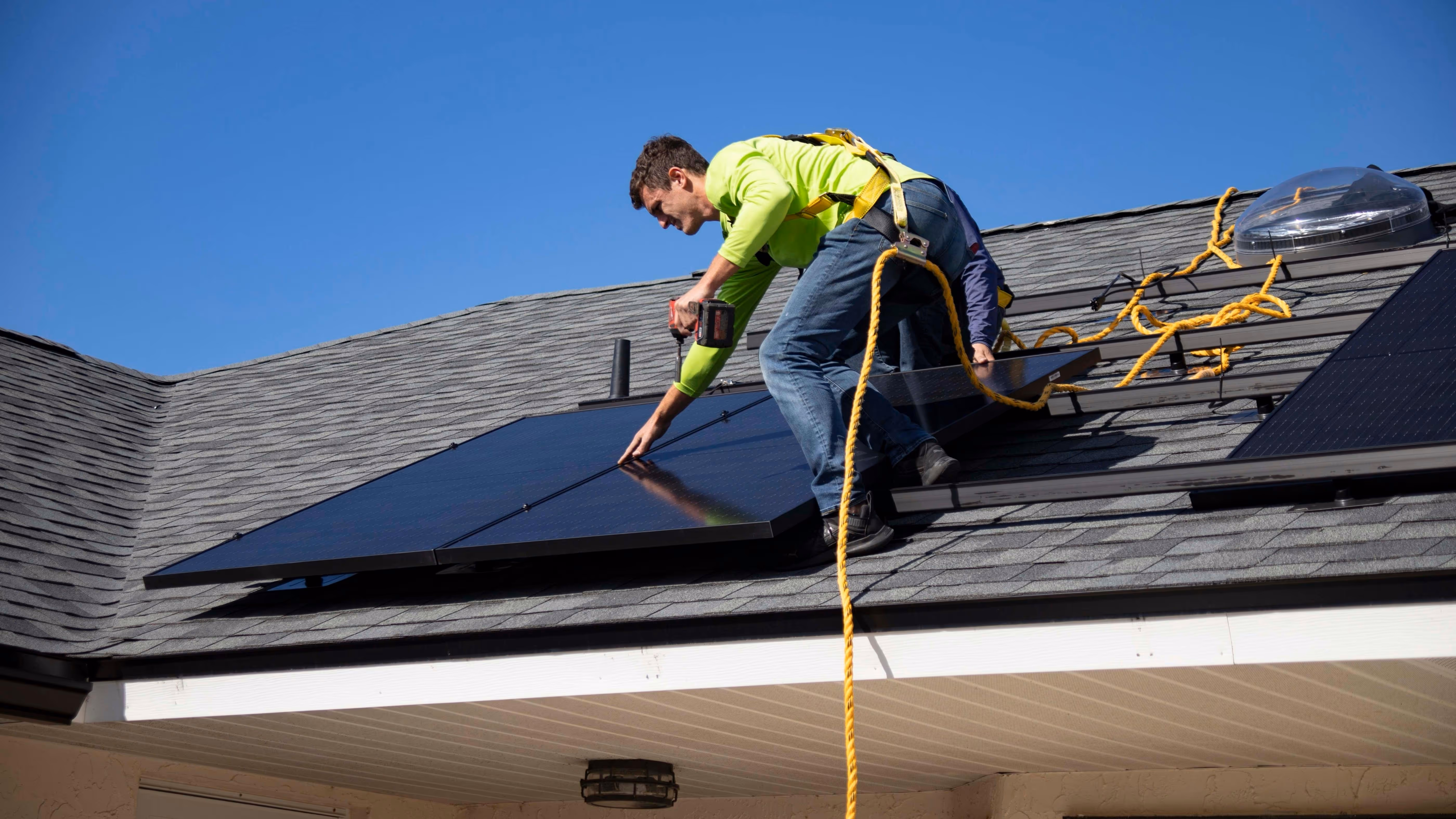A worker in a green shirt and safety harness installs solar panels on a house roof under a clear blue sky.
