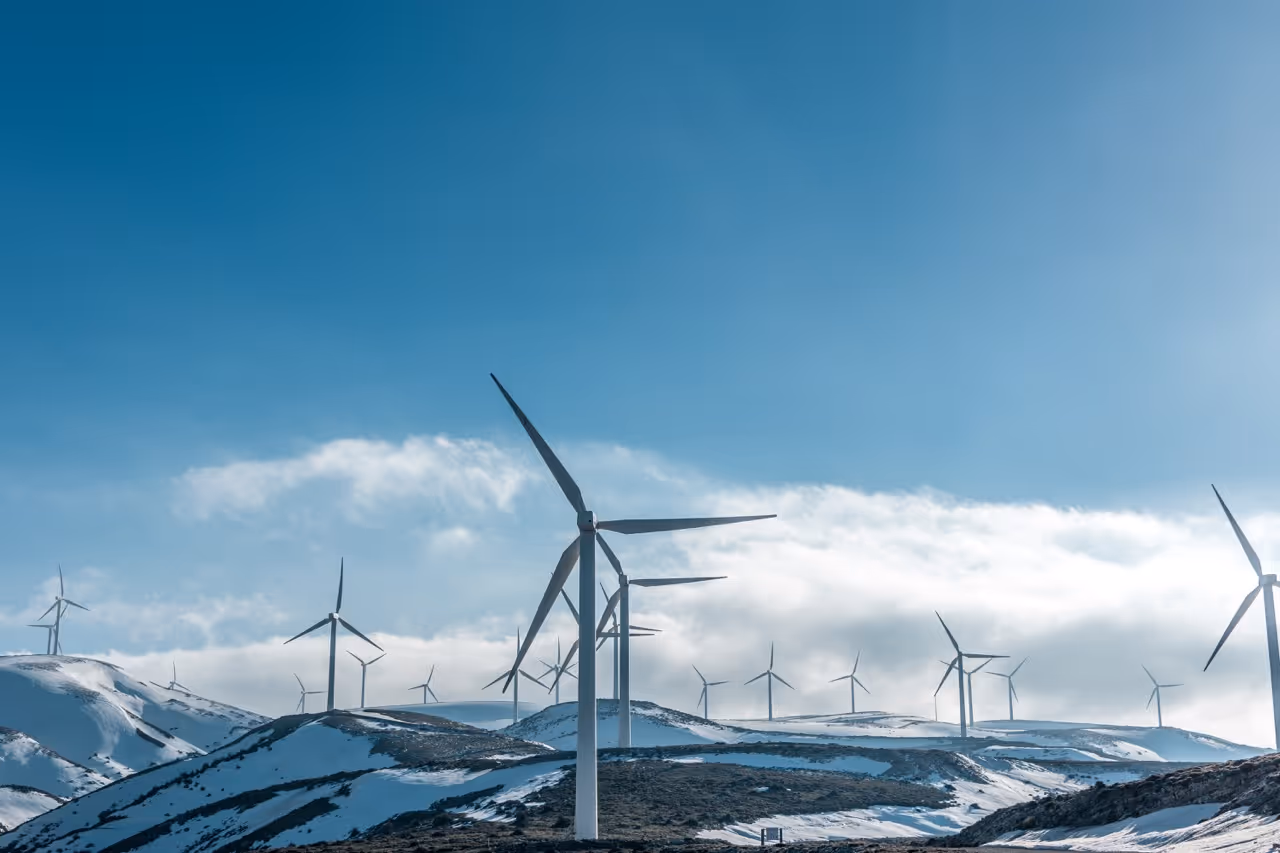 Multiple wind turbines on snow-covered hills under a blue sky with scattered clouds.