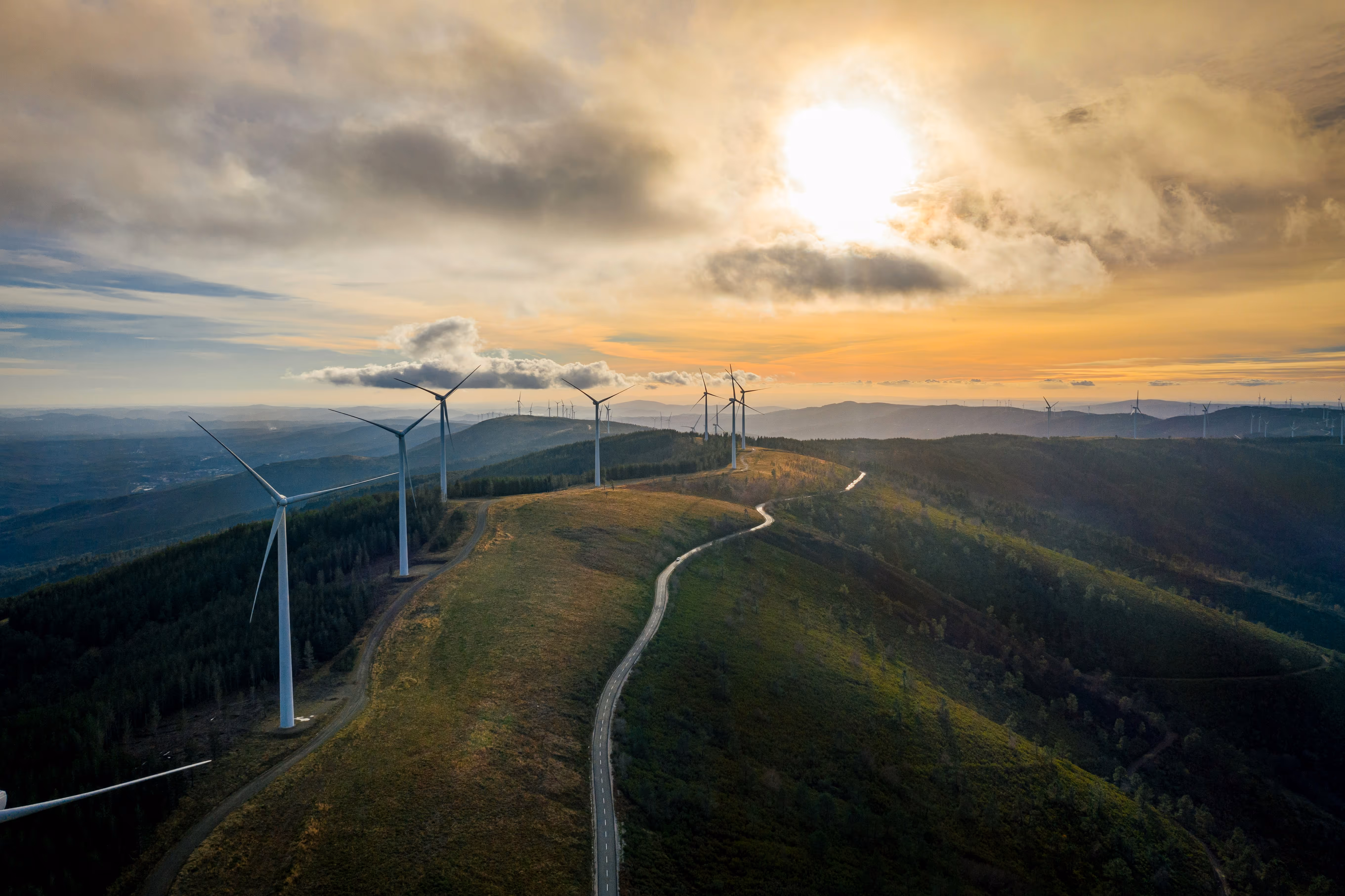Wind turbines on a ridge with a winding road under a partly cloudy sky at sunset.